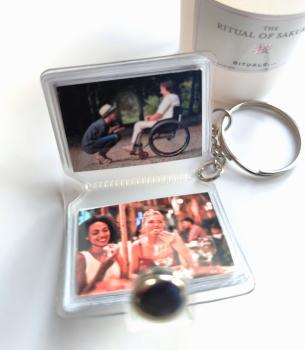 Schlüsselanhänger mit Mini-Fotobuch, das zwei Bilder enthält: eine Person im Rollstuhl im Gespräch mit einer anderen Person im Freien und zwei Frauen, die in einem Restaurant essen und lachen. Im Hintergrund ist eine Rituals-Kerze zu sehen.